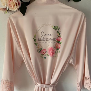 May include: A pale pink satin robe with a floral wreath design and the text "Emma Bridesmaid 1st August 2020". The robe has lace trim on the sleeves and a matching sash.