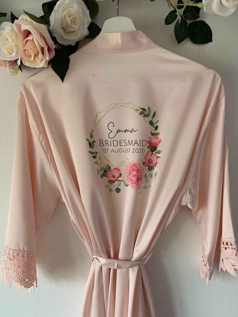 May include: A pale pink satin robe with a floral wreath design and the text "Emma Bridesmaid 1st August 2020". The robe has lace trim on the sleeves and a matching sash.