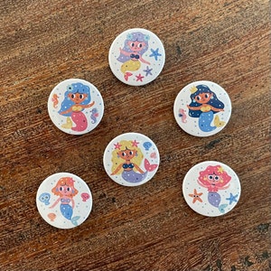 Puede incluir: Seis botones redondos blancos con ilustraciones coloridas de sirenas. Cada botón presenta una sirena diferente con un peinado, una cola y accesorios únicos. Las sirenas están rodeadas de criaturas marinas y otros elementos con temática oceánica.
