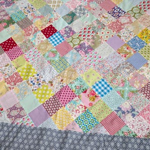 Könnte beinhalten: Ein farbenfrohes Patchwork-Quilt mit einer Vielzahl von gemusterten Stoffen in Rosa-, Grün-, Blau- und Gelbtönen. Der Quilt hat ein geometrisches Design mit einem grau-weißen Sternmuster-Rand und einem rot-weiß karierten Rand.