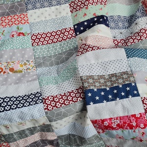 Könnte beinhalten: Eine Patchwork-Decke mit einer Vielzahl von bunten Stoffen, darunter Blumenmuster, geometrische Designs und einfarbige Stoffe. Die Decke besteht aus horizontalen Stoffstreifen, die zusammengenäht sind.