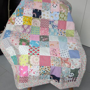 Könnte beinhalten: Eine Patchwork-Decke aus verschiedenen Stoffquadraten mit unterschiedlichen Mustern, darunter Pandas, Regenbögen, Einhörner, Blumen, Punkte und Vichy-Karos. Die Decke ist mit einem floralen Stoff eingefasst und enthält ein kleines rechteckiges Etikett.