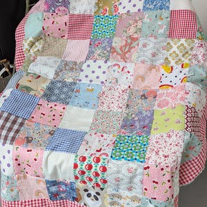 Könnte beinhalten: Eine Patchwork-Decke aus verschiedenen quadratischen Stoffstücken mit diversen Mustern wie Blumen, Vichy-Karos, Punkten und Tiermotiven. Die Decke ist mit einer rot-weiß karierten Bordüre und einem ausgefransten weißen Rand versehen.