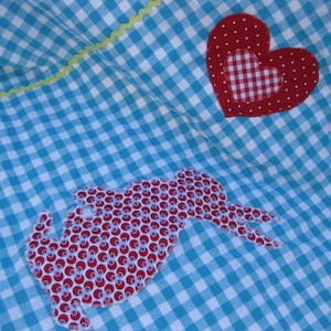Op de afbeelding: Een blauwe en witte gingham stof met een rode hart en een rode konijnenapplicatie. Het hart heeft een wit gingham midden.