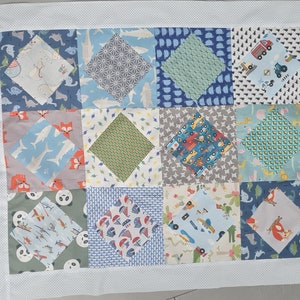 Könnte beinhalten: Ein Patchwork-Quilt mit einer Auswahl an farbenfrohen Stoffquadraten. Die Quadrate zeigen Muster von Dinosauriern, Lastwagen, Füchsen und geometrischen Formen. Der Quilt hat einen weißen Rand mit kleinen, gleichmäßig verteilten Punkten.