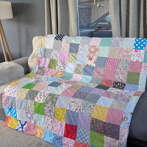 Op de afbeelding: Een patchwork quilt opgebouwd uit talloze vierkante stoffen stukjes in diverse kleuren en patronen, waaronder bloemen-, stippen-, ruitjes- en effen prints. De quilt heeft een rasterindeling met uiteenlopende textielontwerpen.