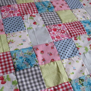 Könnte beinhalten: Ein Patchwork-Quilt mit einer Vielzahl von gemusterten Quadraten. Der Quilt zeigt Quadrate aus rosa, blauen, roten und grünen Stoffen mit Tupfen, Blumendrucken, Vichy-Karo und Fruchtdesigns. Der Quilt ist aus weichem Stoff gefertigt.