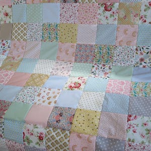 Könnte beinhalten: Ein Patchwork-Quilt in Pastelltönen. Der Quilt besteht aus Quadraten aus rosa, blauen, grünen und gelben Stoffen mit floralen, gepunkteten und geometrischen Mustern. Der Quilt liegt auf einer Oberfläche.