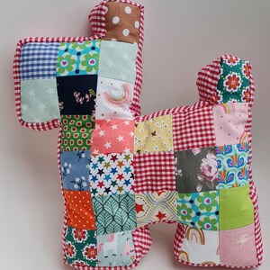 Op de afbeelding: Een patchwork kussen in de vorm van een hond met een rode en witte gingham rand. Het kussen is gemaakt van verschillende kleurrijke stoffen met patronen zoals sterren, bloemen en regenbogen.