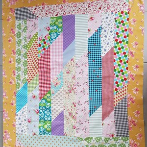 Op de afbeelding: Een kleurrijk quilt met een geometrisch patroon. De quilt is gemaakt van verschillende stoffen met bloemen-, stippen- en streepjespatronen. De quilt is omzoomd met een gele stof met een bloemenpatroon.