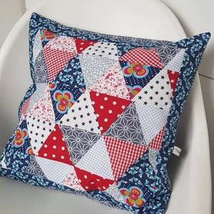 Op de afbeelding: Een blauw, rood en wit patchwork kussen met een geometrisch patroon. Het kussen is gemaakt van verschillende stoffen met verschillende prints, waaronder stippen, sterren en bloemen.