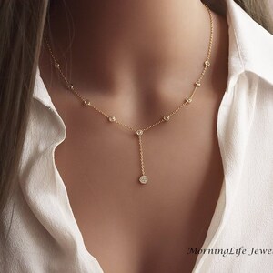 May include: A delicate gold necklace with small, round, clear gemstones. The necklace has a Y-shaped design, with a single gemstone pendant at the end. The necklace is worn against a white shirt. The text "MorningLife Jewelry" is visible.