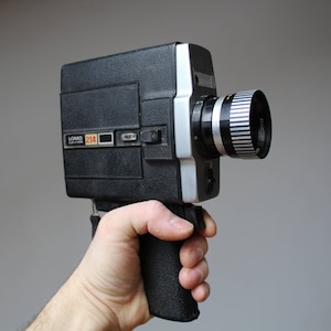 May include: Black and silver Lomo 214 vintage film camera with a textured grip. The camera has a prominent lens with silver and black stripes. The camera is held in a hand against a gray background.