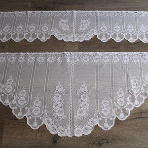 May include: Two white lace window valances with a floral design. The valances have a scalloped edge and a repeating pattern of flowers and leaves. The lace is delicate and airy, suitable for a window.