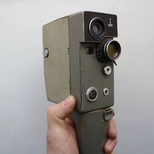 May include: Vintage olive green film camera with a black top section. The camera has a lens, dials, and a handle. The camera is held by a hand against a white background. The camera appears to be in good condition.
