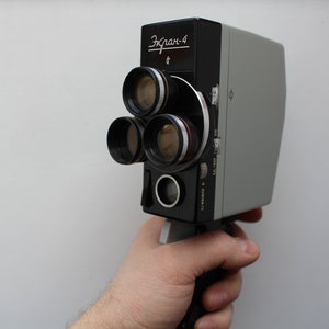 Puede incluir: Una cámara de cine vintage Ekran-4, sostenida por una mano. La cámara tiene un cuerpo negro y gris con tres lentes y el texto "Ekran-4" en escritura cirílica. Las lentes de la cámara tienen detalles en rojo y amarillo.
