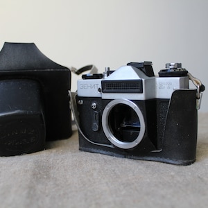 Puede incluir: Una cámara de película Zenit ET de 35 mm plateada y negra vintage con una funda de cuero. La cámara tiene una montura de lente, un visor y varios diales y botones. La funda de cuero negro está al lado de la cámara.