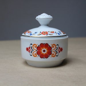 Op de afbeelding: Witte keramische suikerpot met deksel, versierd met een bloemmotief in rood, oranje en zwart. Het deksel heeft een kleine handgreep. De rand van het deksel is afgewerkt met goud.