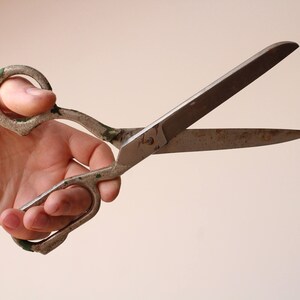May include: A pair of heavy-duty metal shears with a weathered, silver-toned finish and visible signs of oxidation. The handles feature remnants of green paint, and the long, pointed blades show surface wear and discoloration from age and use.