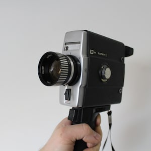 May include: A vintage Super 8 film camera with a black and silver body and a large lens. The camera has a handle and a wrist strap. The lens is black with silver rings. The camera is held against a white background.