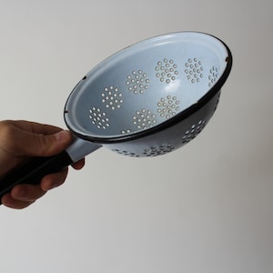 May include: A light blue enamel colander with a black handle and rim. The colander has numerous small holes for draining liquids. The handle is black and has a hole at the end for hanging. A kitchen utensil.