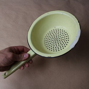 Könnte beinhalten: Ein blassgelbes Emaille-Sieb mit einem langen Griff, der am Ende ein Loch zum Aufhängen hat. Die Schüssel verfügt über einen perforierten Boden zum Abtropfen und einen schwarzen Rand mit sichtbaren weißen Emaille-Abplatzungen am Beckenrand.