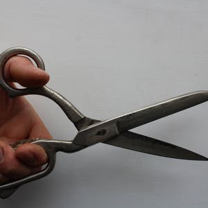 May include: A pair of heavy-duty metal shears with a weathered, silver-toned finish. The tool features two large, circular finger loops and long, pointed blades that are partially open, showing signs of age and use on the metallic surface.