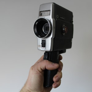 May include: A vintage Super 8 film camera with a silver and black body. The camera has a lens, a viewfinder, and a handle. The word "VHS" is printed on the front of the camera.