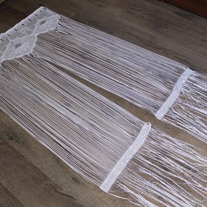 May include: A white lace curtain panel featuring an intricate floral pattern at the top header, followed by long, flowing vertical fringe strands that extend downward, ending in a secondary horizontal band with a geometric diamond design.