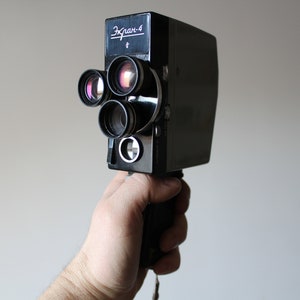 Puede incluir: Cámara de cine vintage Ekran-4 negra con múltiples lentes. La cámara tiene un cuerpo rectangular y un asa. El texto "Ekran-4" es visible en la parte superior de la cámara.