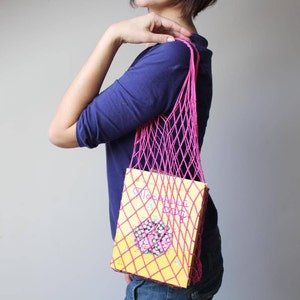 May include: A bright pink string mesh bag with a diamond-patterned weave. The bag contains a rectangular yellow book with Cyrillic text and a graphic illustration of a flower on the front cover. The bag features long straps for shoulder carrying.
