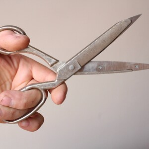 May include: A pair of silver-toned metal scissors with two finger loops and pointed blades. The metal surface shows a weathered, matte texture with visible signs of wear, including small scratches and a central pivot screw connecting the two blades.
