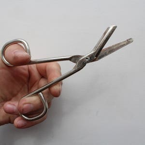 May include: A pair of silver-toned metal bandage scissors with a blunt, angled tip on the lower blade and two rounded finger loops. The scissors feature a metallic finish with visible signs of wear and a central pivot screw connecting the two blades.