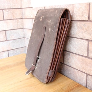 May include: A vintage brown leather document case with a buckle closure and a strap. The case has a worn appearance, suggesting age and use. The interior appears to have multiple compartments for organizing papers.
