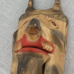 May include: A carved wooden figure with a stylized face, featuring a prominent nose, red lips, and dark accents. The figure has two upward-pointing projections and is painted in shades of brown and tan.