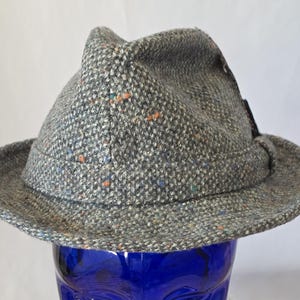 May include: A gray tweed fedora hat with a slightly upturned brim. The hat features a textured fabric with flecks of blue, orange, and green. The hat is set against a blue background.