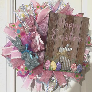 May include: A pink and blue Easter wreath with a wooden sign that says "Happy Easter". The wreath is decorated with pink and blue ribbons, glitter bunnies, and Easter eggs.