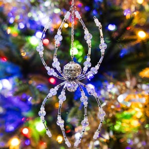 Puede incluir: Adorno navideño de araña con cuerpo plateado y patas transparentes con cuentas. La araña está suspendida por una cadena plateada, con un fondo borroso de luces de árbol de Navidad.