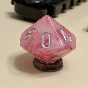 以下が含まれることがあります： 銀色の数字が書かれたピンクと白の大理石模様のd8サイコロ。サイコロは茶色の台座の上に置かれています。