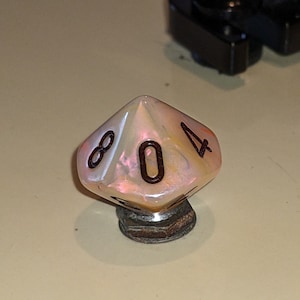Può includere: Un dado d10 bianco e arancione iridescente con numeri neri, che mostra il numero 0. Il dado è appoggiato su una base di metallo.