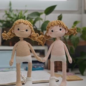 May include: Two handmade crochet dolls with golden-brown yarn hair styled in pigtails. One doll has a tan complexion, the other a light pink complexion. Both dolls wear white crocheted underwear and a single white sock on one foot.
