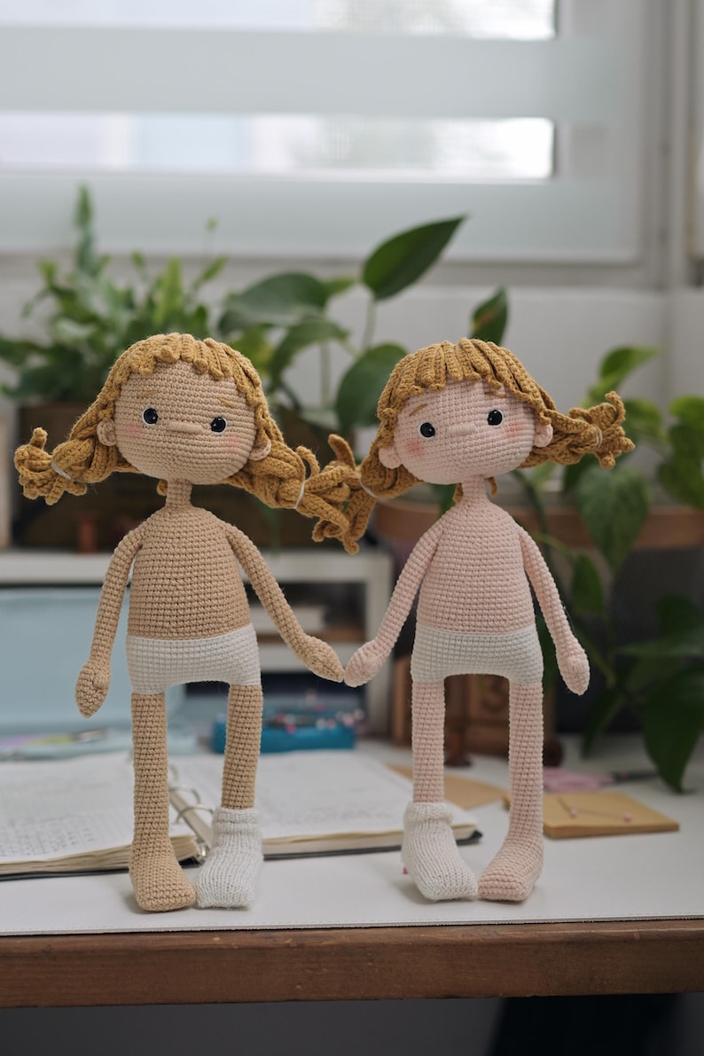 May include: Two handmade crochet dolls with golden-brown yarn hair styled in pigtails. One doll has a tan complexion, the other a light pink complexion. Both dolls wear white crocheted underwear and a single white sock on one foot.