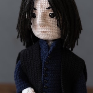 May include: Crocheted doll with black hair, wearing a black jacket over a blue vest with black buttons. The doll has a white face with black eyes and a serious expression.
