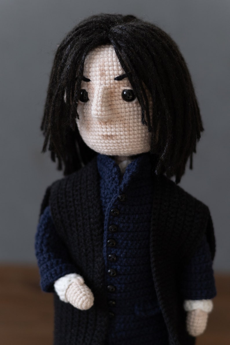May include: Crocheted doll with black hair, wearing a black jacket over a blue vest with black buttons. The doll has a white face with black eyes and a serious expression.