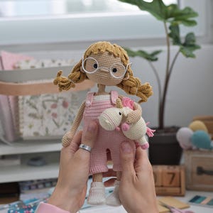 May include: A handmade crochet doll with blonde yarn hair styled in pigtails, wearing white round glasses and pink overalls. The doll is holding a small white and pink crochet unicorn toy. The doll has a light beige complexion and white socks.