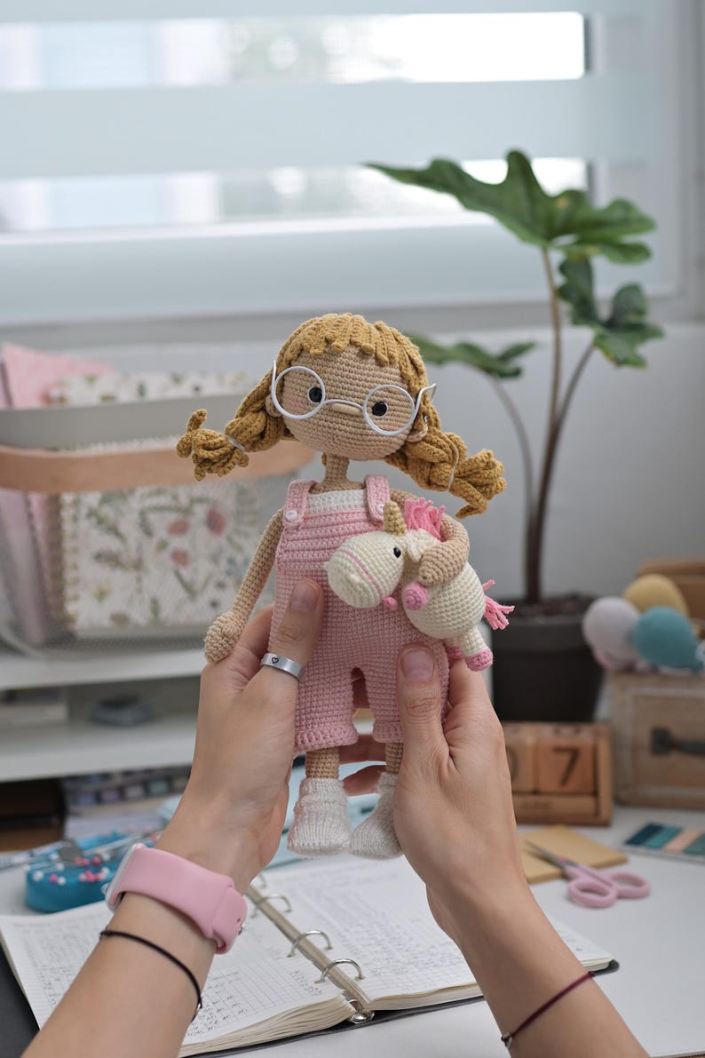May include: A handmade crochet doll with blonde yarn hair styled in pigtails, wearing white round glasses and pink overalls. The doll is holding a small white and pink crochet unicorn toy. The doll has a light beige complexion and white socks.