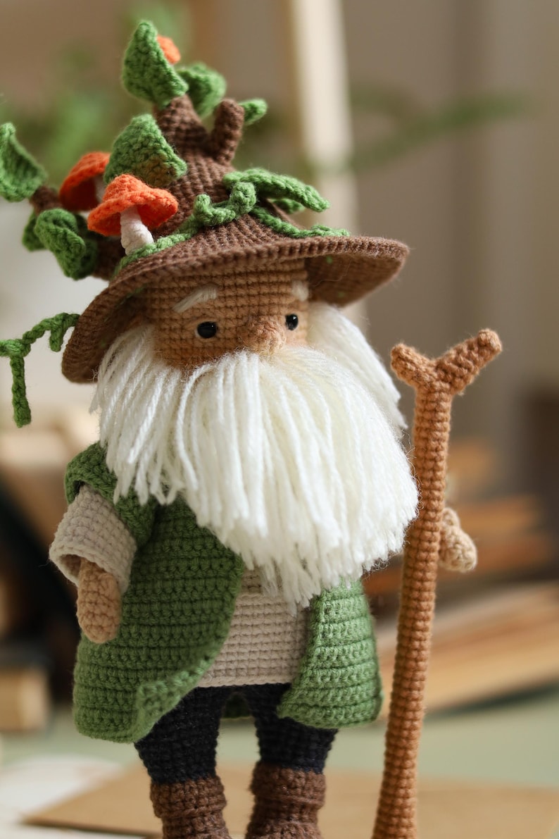 May include: A crocheted gnome with a brown hat topped with green leaves and orange mushrooms. The gnome is wearing a green jacket, brown pants, and brown boots. The gnome is holding a brown stick.