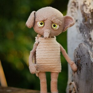 Puede incluir: Un juguete de peluche de crochet de un elfo dom&eacute;stico con orejas grandes, una nariz larga y ojos verdes. El elfo lleva una t&uacute;nica marr&oacute;n simple y est&aacute; de pie sobre una superficie de madera.