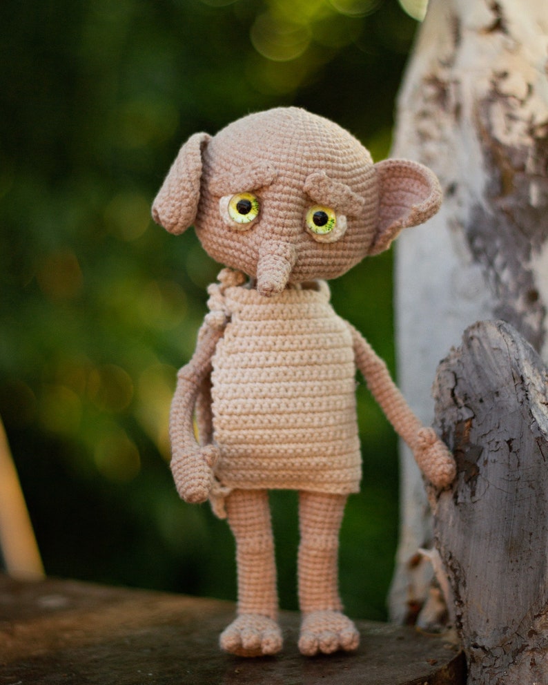 Puede incluir: Un juguete de peluche de crochet de un elfo dom&eacute;stico con orejas grandes, una nariz larga y ojos verdes. El elfo lleva una t&uacute;nica marr&oacute;n simple y est&aacute; de pie sobre una superficie de madera.