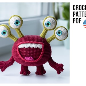 May include: Crochet pattern PDF for a red, three-eyed monster with a big smile and green antennae. The monster has blue eyes and is standing on a white surface.